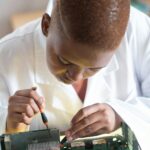 Focused technician in lab coat checking current of electricity with voltmeter while examining condition of graphics card in workshop